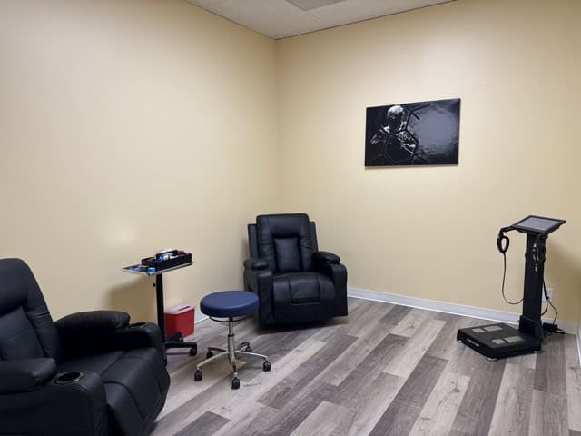 A small medical office with beige walls, two black reclining chairs, a blue stool, a side table with medical supplies, a scale with a digital screen, and a black-and-white photo on the wall.