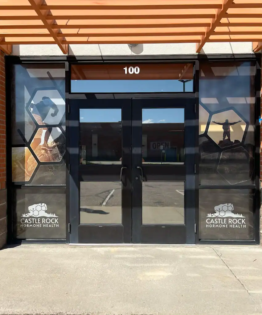 A glass door entrance to Castle Rock Hormone Health in Fort Collins features the number 100 above the door and hexagonal designs on the windows. The logo and name appear at the bottom of both side windows.