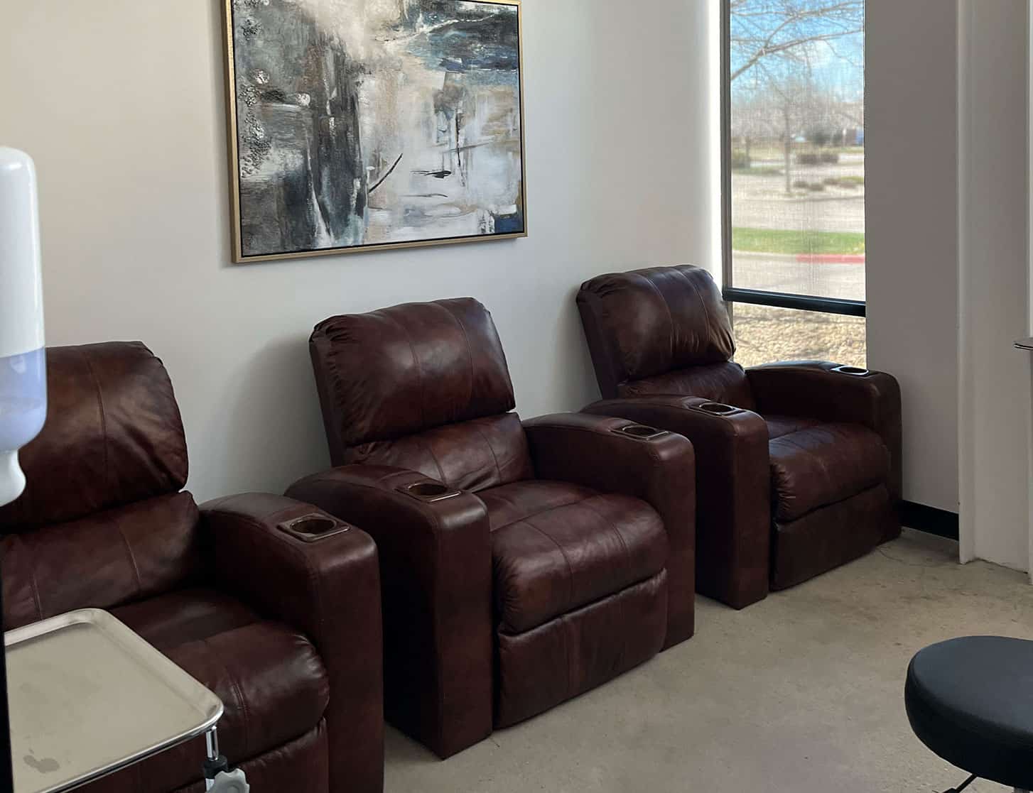 Three brown leather recliners are lined up against a white wall below an abstract painting, next to a large window with a view of trees and a parking lot outside.