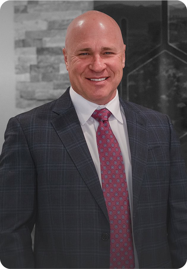 A bald man in a plaid suit and pink patterned tie smiles while standing indoors against a modern, neutral-toned background.