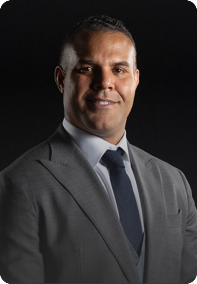 A man in a gray suit, white shirt, and dark tie is smiling against a plain black background.