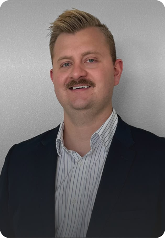 A man with short blond hair and a mustache, wearing a dark suit jacket over a white striped shirt, smiles while standing in front of a plain light gray background.