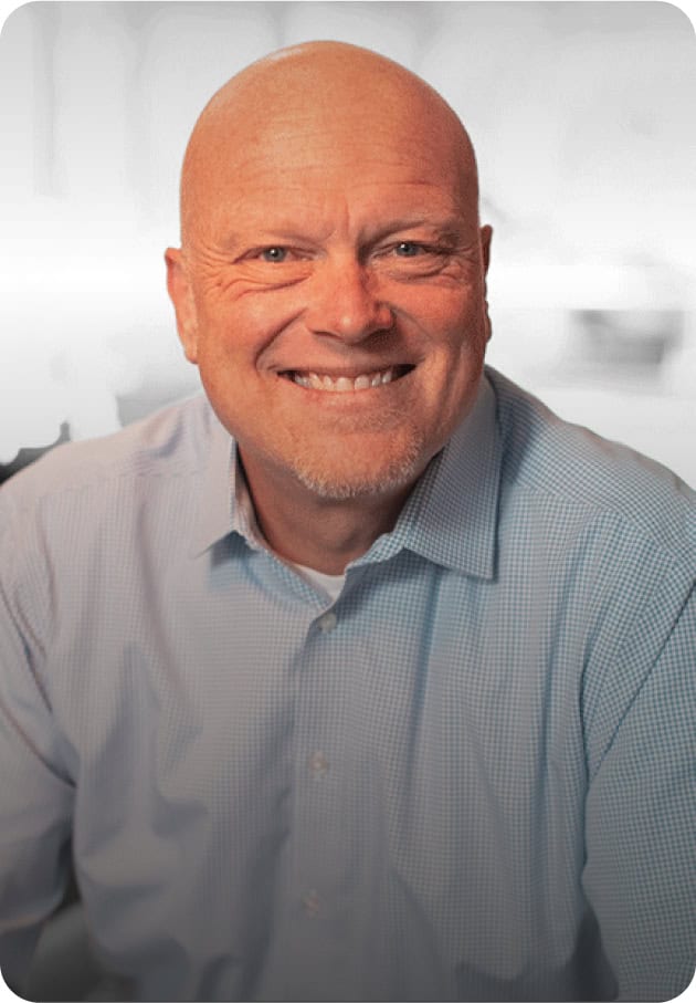 A smiling older man with a bald head and light goatee, wearing a light blue checkered shirt, sits facing the camera against a softly blurred background.
