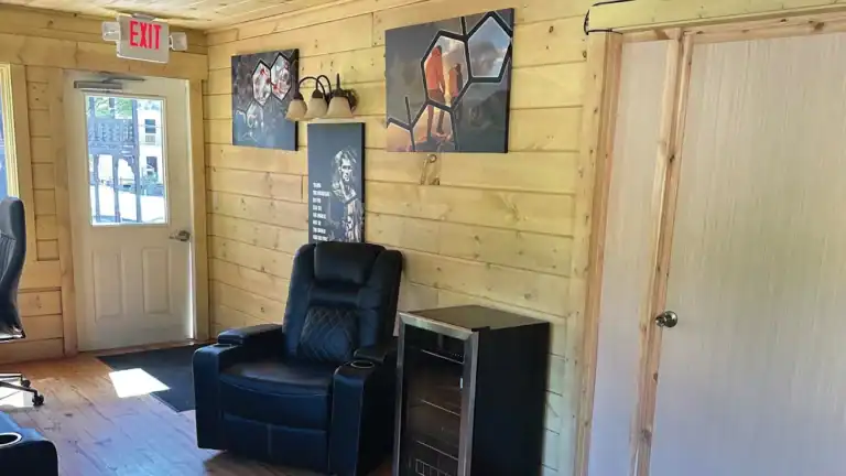A black recliner sits in a wood-paneled room with a door, wall art, and a small shelf nearby. Sunlight enters through a window next to the door, creating a cozy atmosphere.