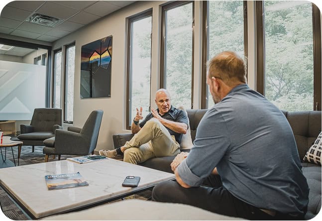 Two men sit on sofas in a modern office lounge, engaged in a serious conversation. One gestures with his hands while the other listens. Large windows let in daylight, and magazines rest on a coffee table between them.