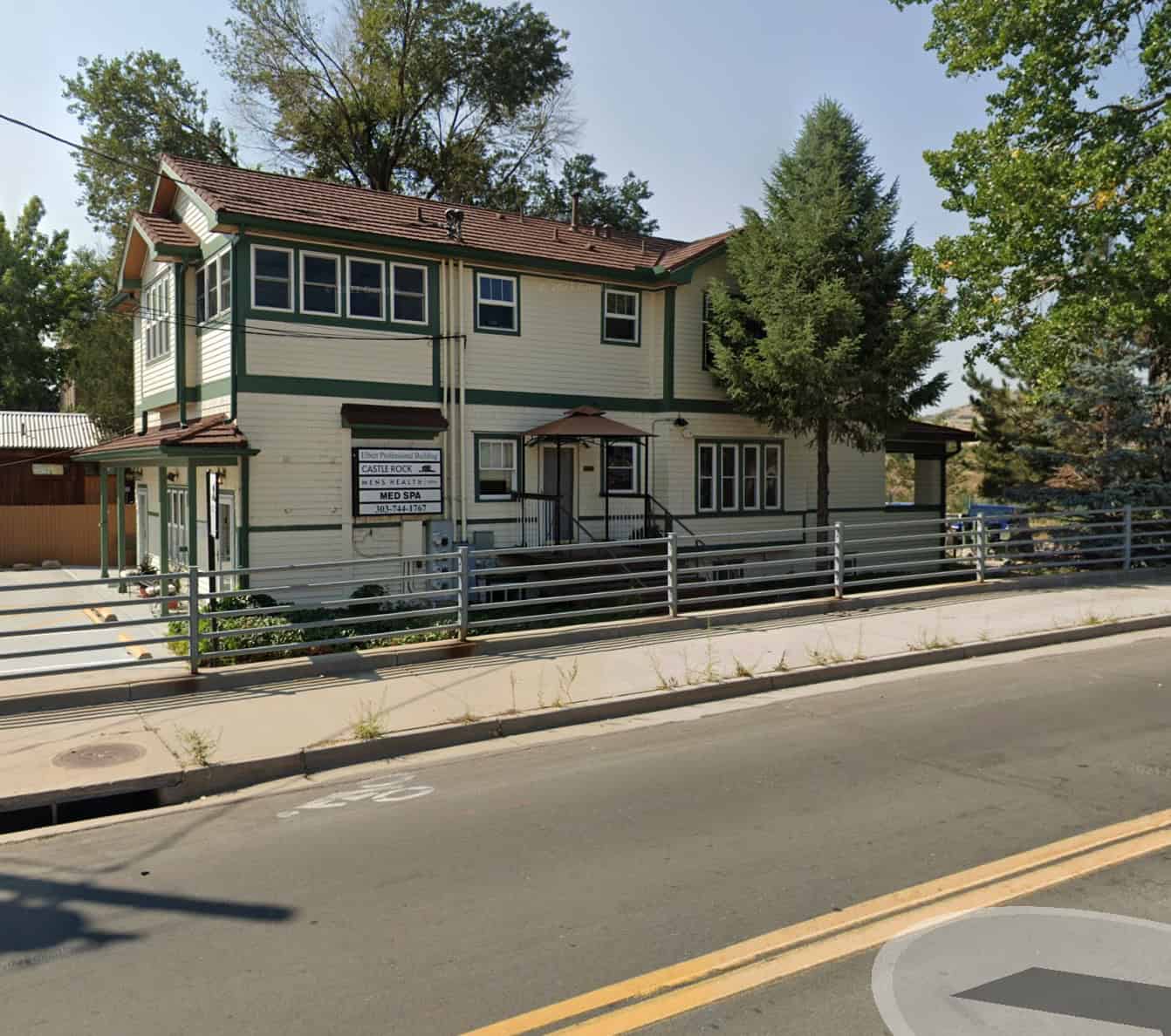 Two-story cream-colored building with green trim and a brown roof, surrounded by trees. A metal fence and sidewalk run along the front, with street and bike lane markings visible in the foreground.