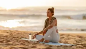 A woman, focused on hormone care, sits cross-legged on a yoga mat on a sandy beach at sunrise or sunset, with a towel draped over her shoulders and a water bottle beside her. The ocean and soft sunlight are in the background.