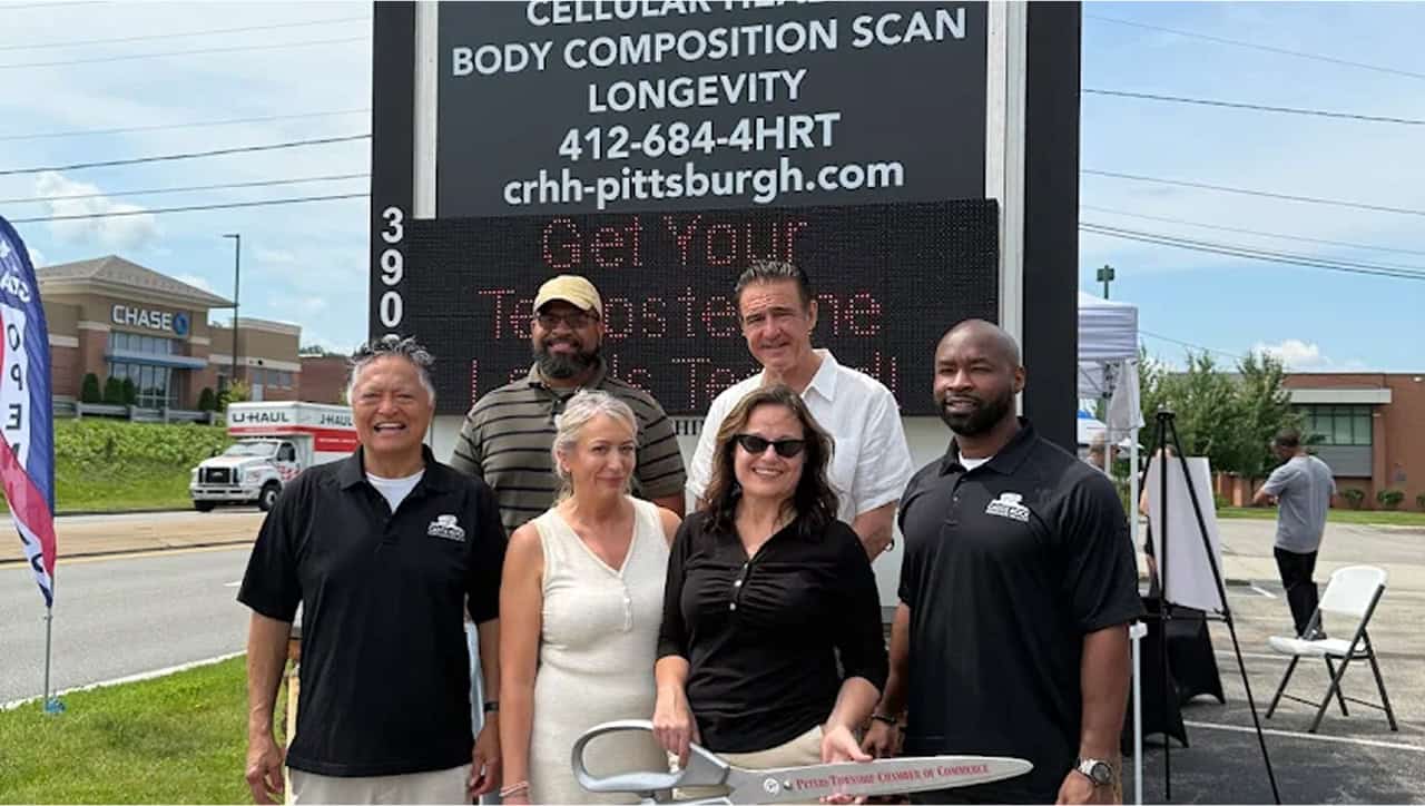 Six people stand smiling in front of a digital sign for a cellular health and longevity center. One woman holds an oversized ceremonial scissors, suggesting a ribbon-cutting event. Buildings, cars, and a tent are visible in the background.