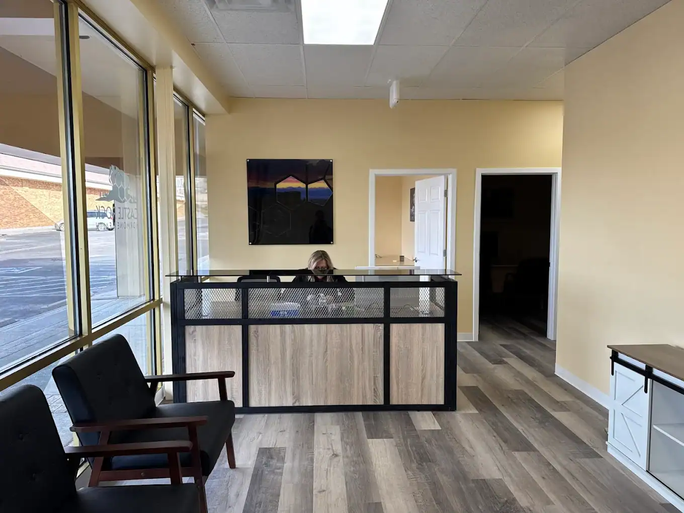 A woman sits behind a modern reception desk in a bright office lobby with yellow walls, large windows, black chairs, and wood-style flooring.