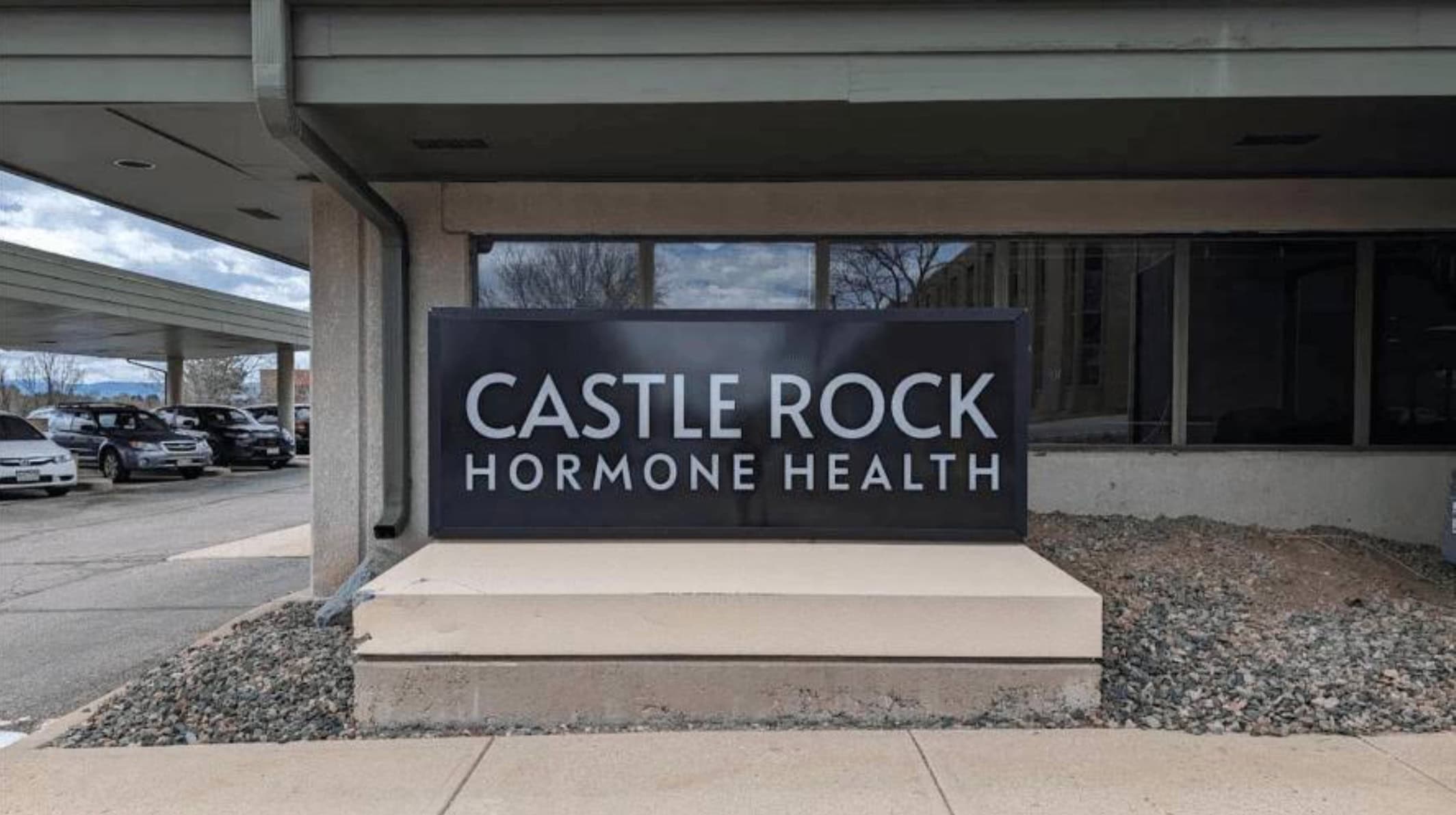 A large black sign with white text reads Castle Rock Hormone Health outside a building, near a parking lot with several cars.