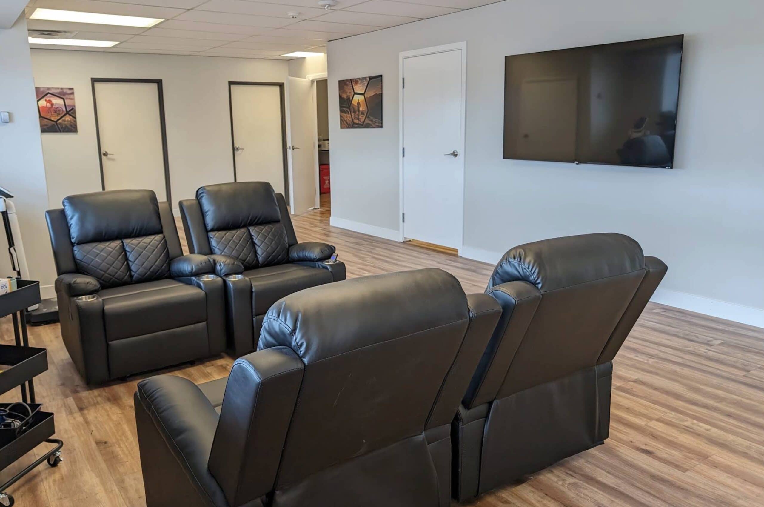 A modern lounge with four black leather recliners facing a wall-mounted flat-screen TV. The room has wood flooring, light walls, three closed doors, and two framed pictures on the walls.