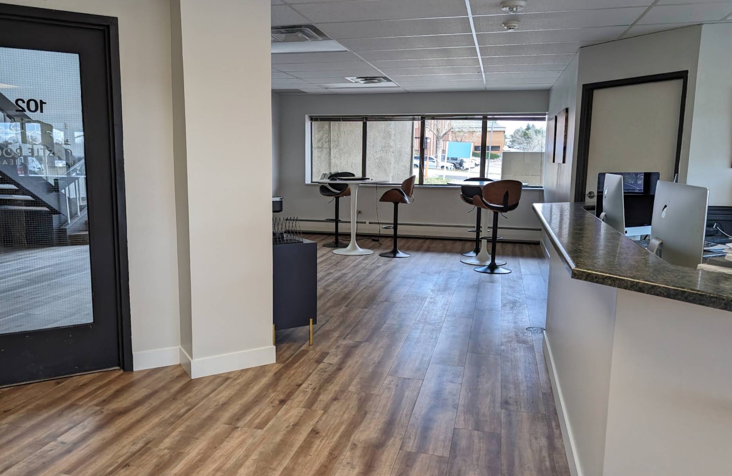 Modern office space with wood flooring, a granite reception counter, barstool chairs by large windows, and computers on desks. Natural light fills the room, creating a clean and open atmosphere.