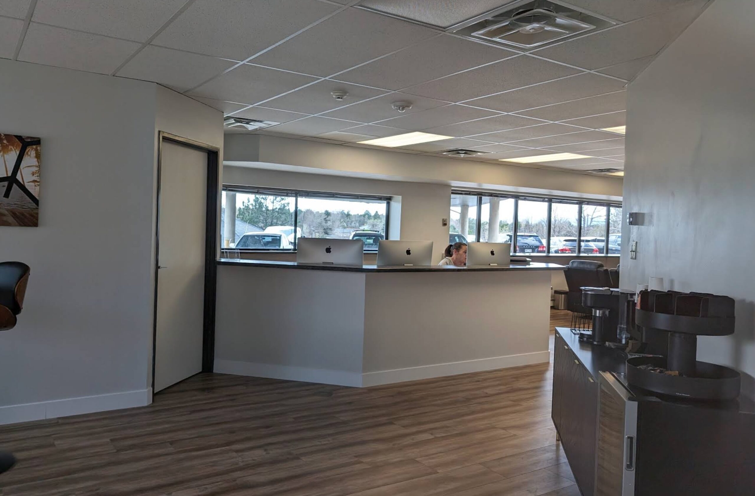 A modern office space with large windows, several computers on a counter, a person working at a desk, wood flooring, and some furniture and office equipment in the foreground.