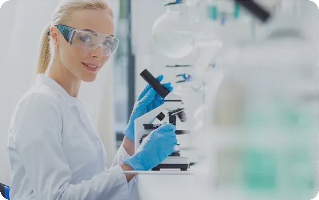 A scientist wearing safety glasses, gloves, and a lab coat looks at the camera while using a microscope in a bright laboratory setting. Laboratory glassware is visible in the background.