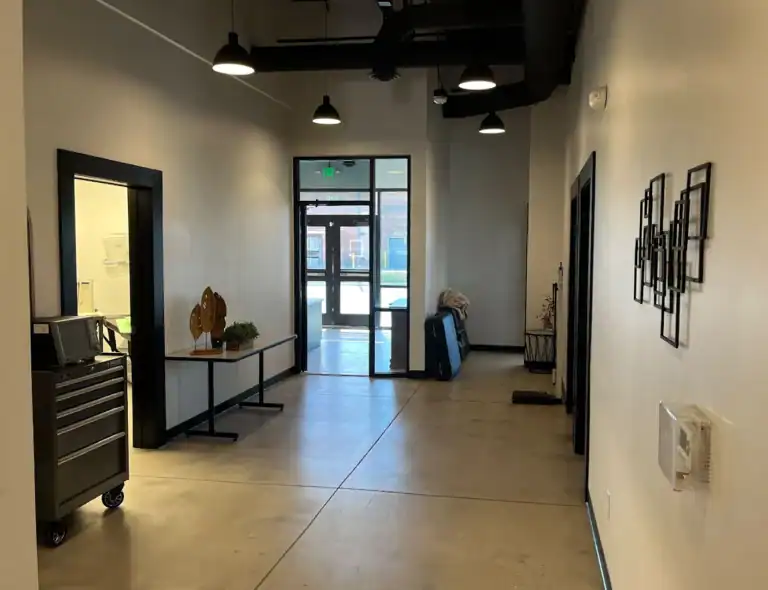 A clean, modern hallway with polished concrete floors, white walls, black trim, and ceiling lights. A table with decor and a rolling cart are on the left, and artwork hangs on the right wall near a glass exit door.
