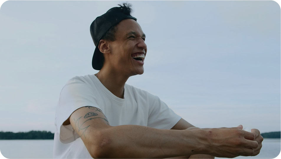 A young person in a white t-shirt and backwards black cap sits outdoors, smiling and laughing. Visible tattoos are on their arm, and a calm body of water with a tree line is in the background.