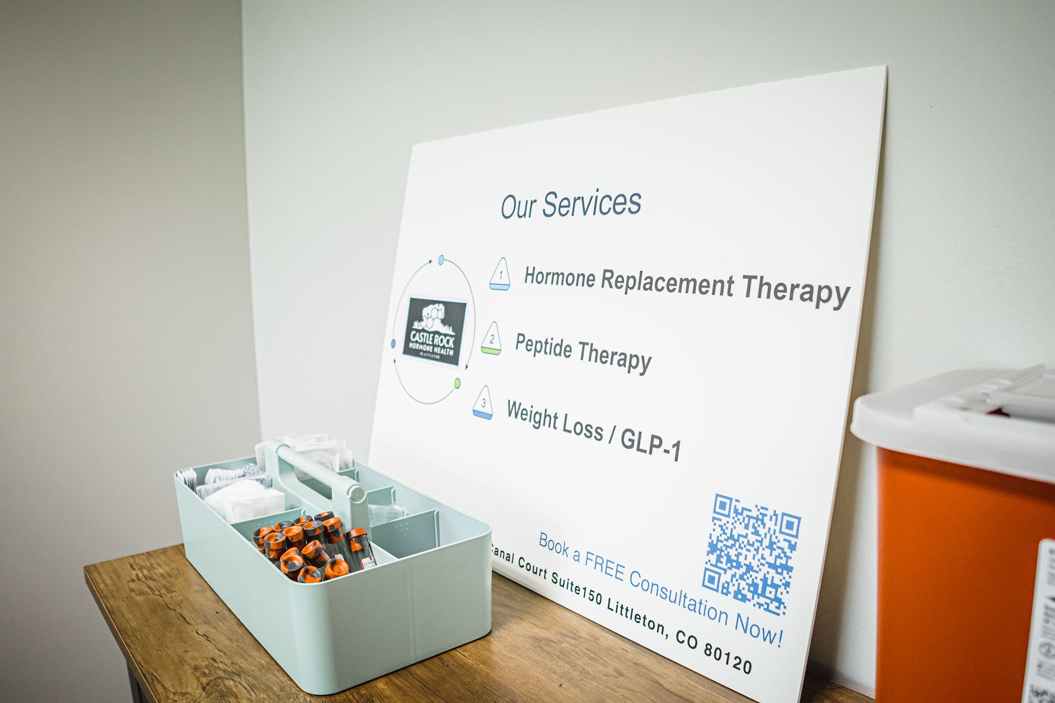 A sign on a table lists medical services: Hormone Replacement Therapy, Peptide Therapy, and Weight Loss/GLP-1. Next to it is a tray with syringes and medical supplies. A sharps container is partially visible.