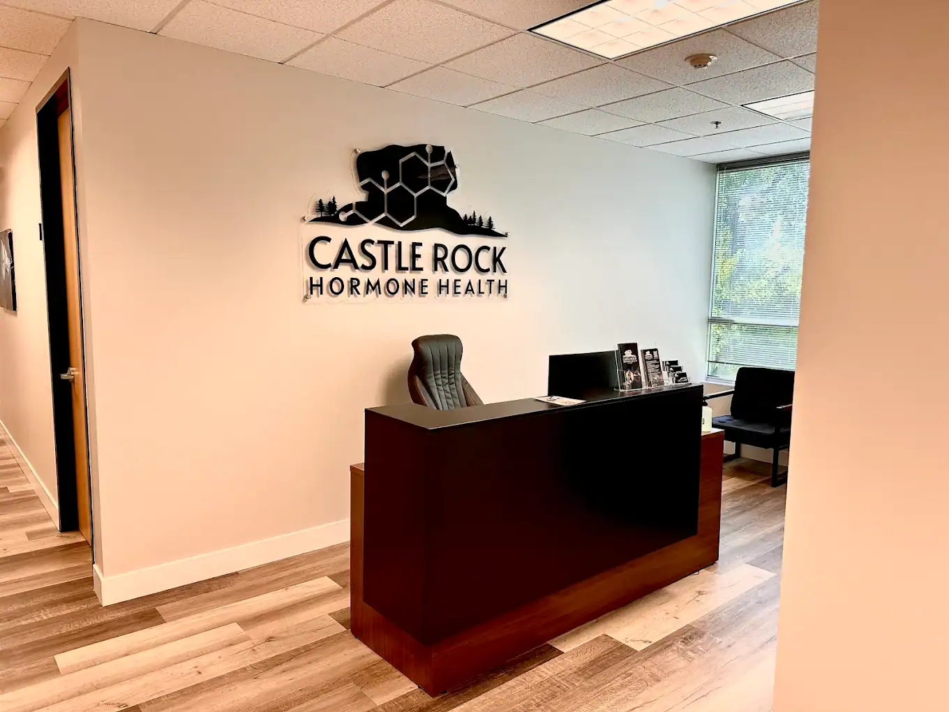 A modern medical office reception area with a dark wood desk, an empty black chair, and a wall sign reading Castle Rock Hormone Health. Natural light enters through a window, and chairs sit in a waiting area.