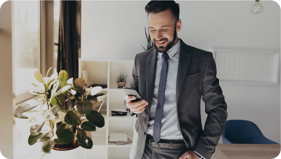 A man in a gray suit stands in a modern office, smiling while looking at his smartphone. There are plants and shelves with decorative items in the background.