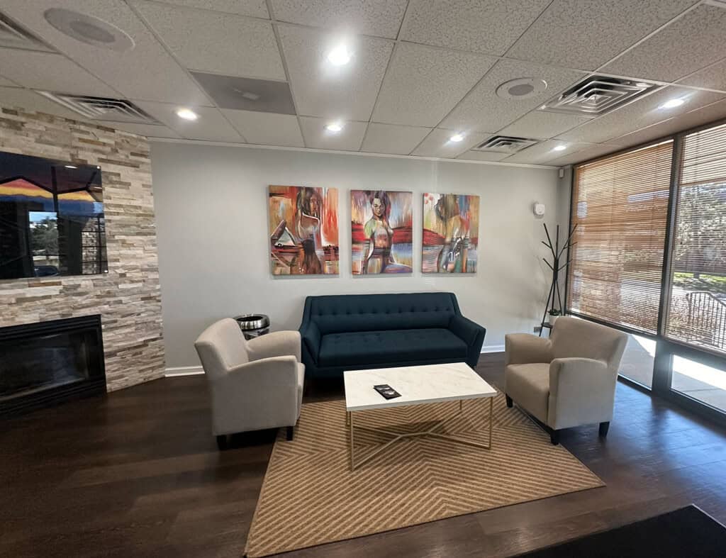 A modern lounge area with a blue sofa, two beige armchairs, a white marble coffee table, and abstract art on the wall; large windows let in natural light and a fireplace is on the left.