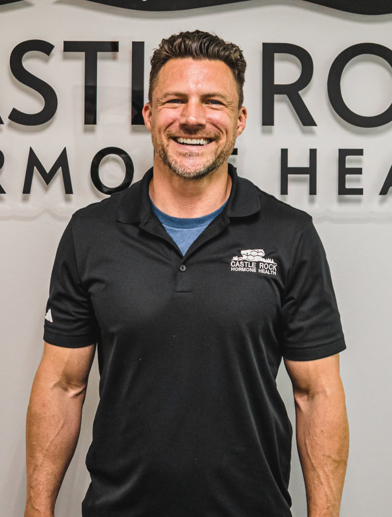 A smiling man with short brown hair, wearing a black polo shirt with the logo Castle Rock Hormone Health, stands in front of a wall with the company’s name displayed in large letters.