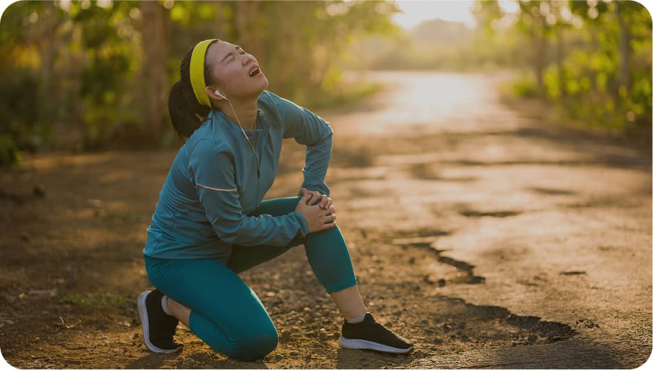 A woman in athletic wear kneels on a sunlit outdoor path, clutching her knee in pain, with a distressed expression on her face. She appears to be injured while exercising or running.