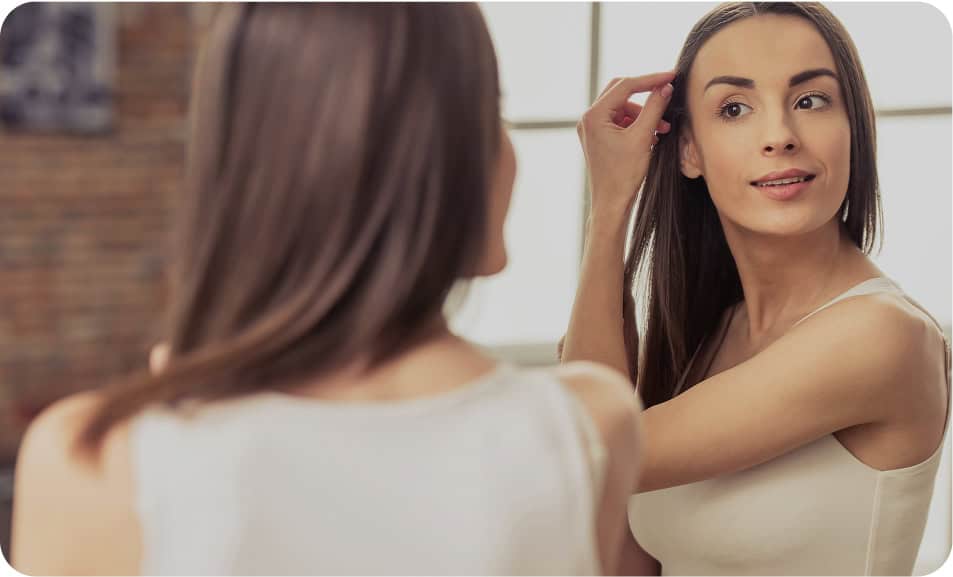 A woman with long brown hair looks at herself in the mirror, adjusting her hair with one hand. She is wearing a light-colored tank top and standing in a softly lit room with a blurred background.