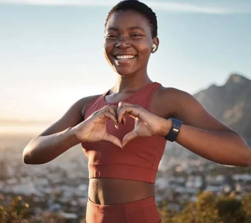 A smiling woman in athletic wear forms a heart shape with her hands outdoors, with a scenic view of mountains and a city in the background. She appears happy and energetic, possibly after exercising.