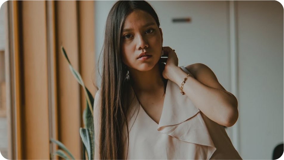 A young woman with long straight hair wearing a sleeveless beige top stands indoors, looking at the camera with a neutral expression and her hand resting on her neck.